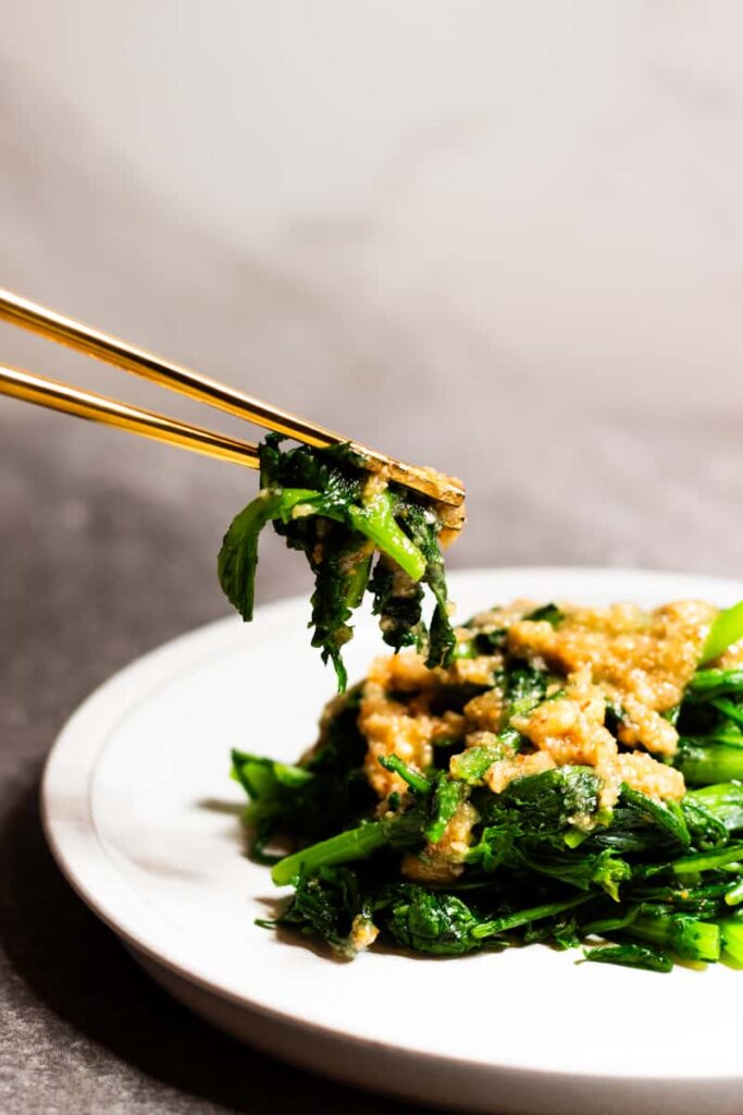 A Pair of Golden Chopsticks Picking Up Some Chrysanthemum Greens with Nutty Sauce on a White Plate Against Grey Background