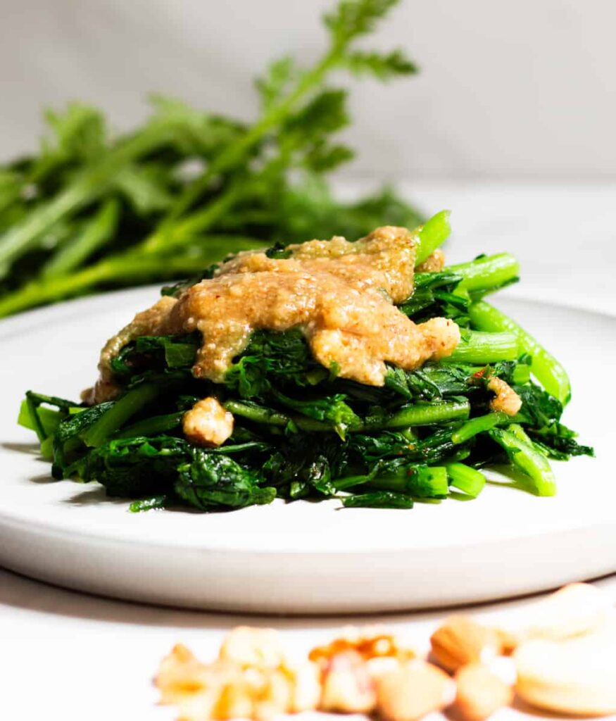 Chrysanthemum Greens Topped with Nut Sauce on a White Plate Against Grey Background with Recipe Ingredients