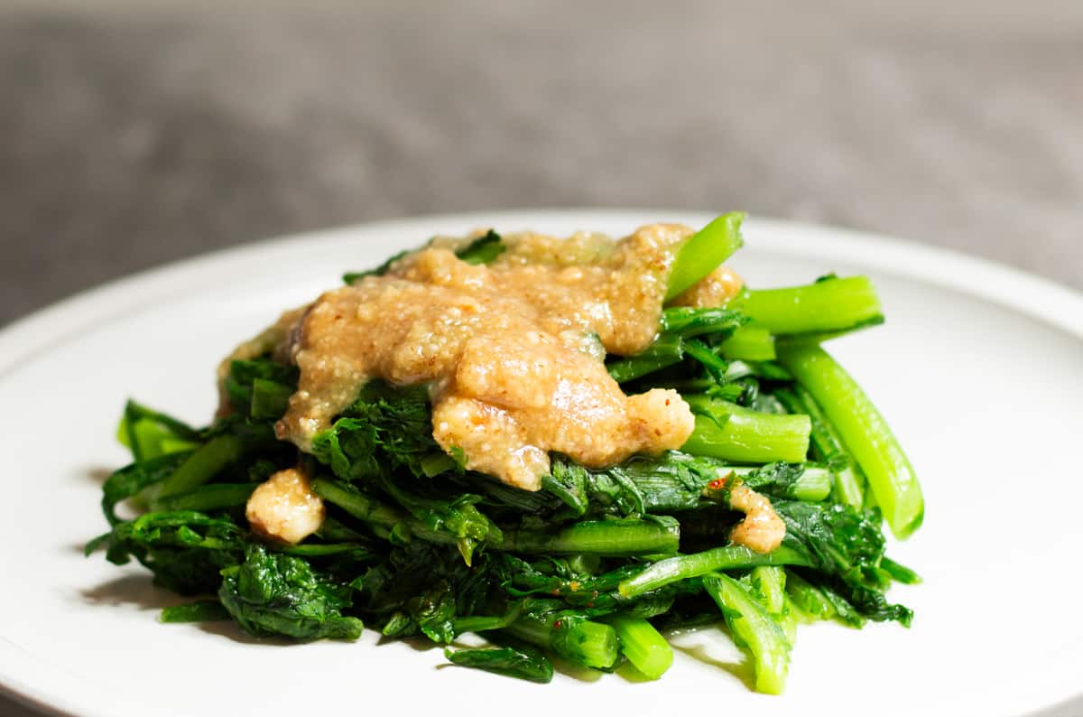 Close Up of Chrysanthemum Greens Topped with Nut Sauce on a White Plate Against Grey Background