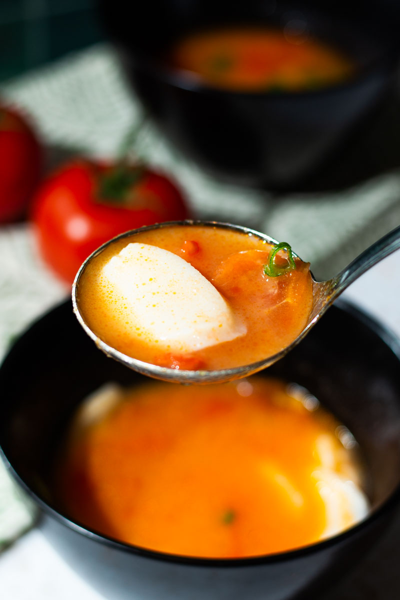 A Ladle of Tomato Rice Cake Soup with Green Onions from A Black Bowl with Tomatoes and Another Bowl of Soup in the Background