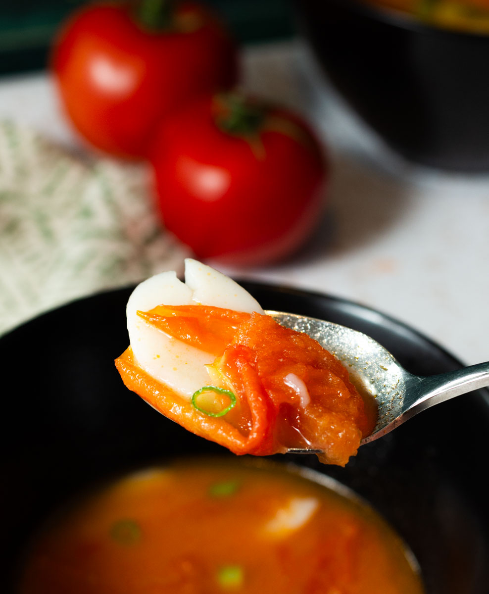 A Spoonful of Tomato Rice Cake Soup with Green Onions from A Black Bowl with Tomatoes and Another Bowl of Soup in the Background