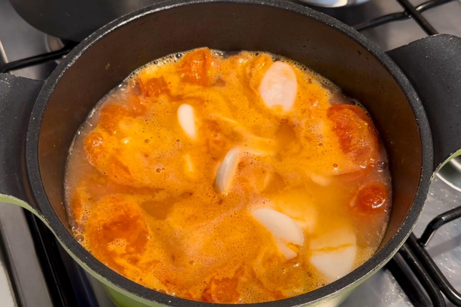 Tomato pieces and sliced rice cakes simmering in broth inside a black pot on the stove.