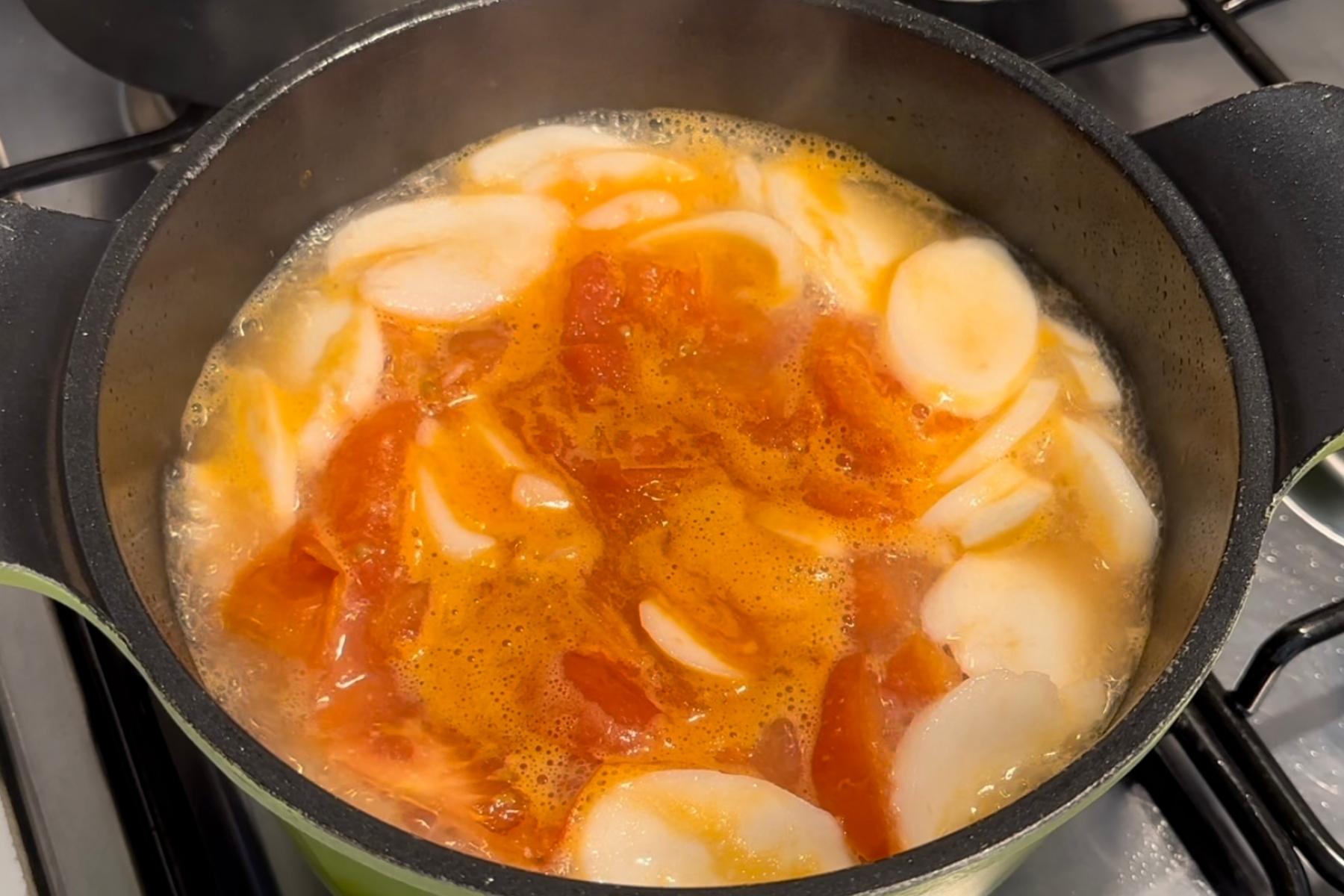 Tomato pieces and sliced rice cakes simmering in broth inside a black pot on the stove.