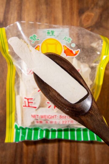 A piece of dried Chinese yam or dried Dioscorea on a dark wooden spoon. There is bag of opened dried Chinese yam on a wooden surface in the background.