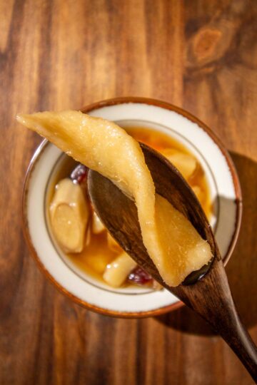 A piece of cooked dried Chinese yam or dried Dioscorea on a dark wooden spoon. There is bowl of burdock root soup on a wooden surface in the background.