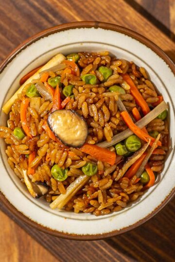 An overhead view of a bowl of burdock root rice, with chopped mushrooms, burdock roots, carrots, and peas visibly incorporated within. There is a pair of wooden chopsticks to its right and they are both sitting on a wooden chopping board.