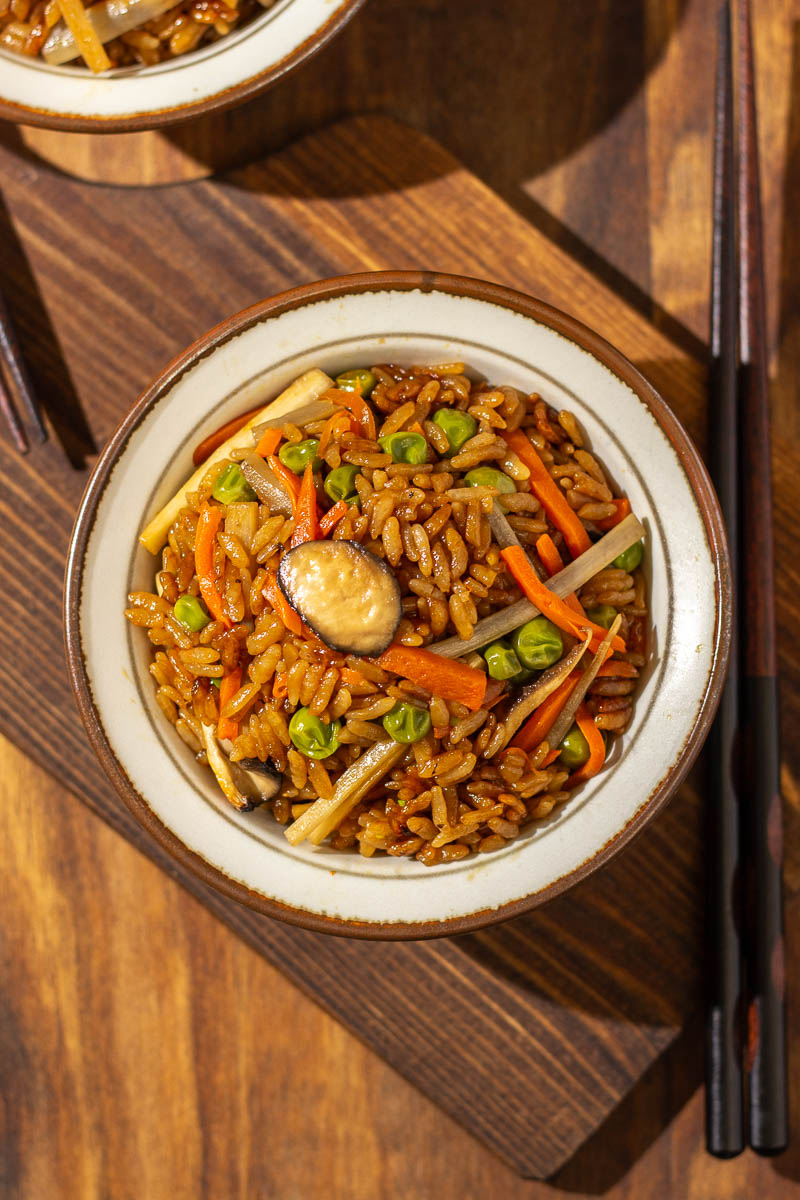An overhead view of a bowl of burdock root rice, with chopped mushrooms, burdock roots, carrots, and peas visibly incorporated within. There is a pair of wooden chopsticks to its right and they are both sitting on a wooden chopping board.
