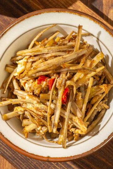 An overhead view of a bowl of burdock root with eggs, with a pair of wooden chopsticks to its right, both sitting on a wooden board and a wooden surface.