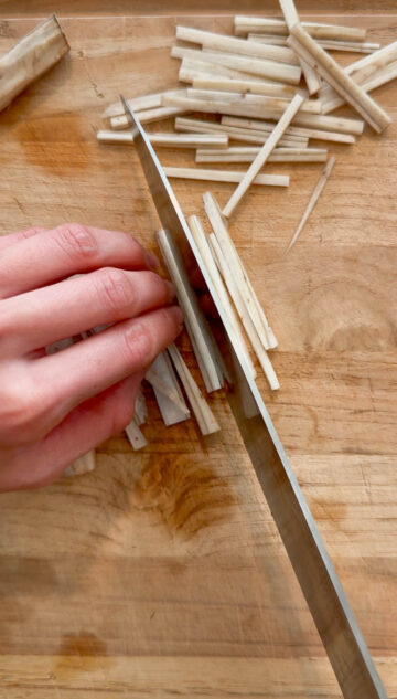 An overhead view of a person julienning burdock roots on a wooden chopping board..
