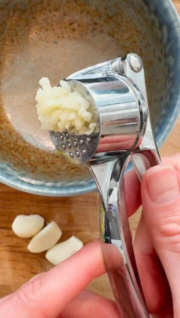 A garlic pressor being used over a bowl on a wooden surface.
