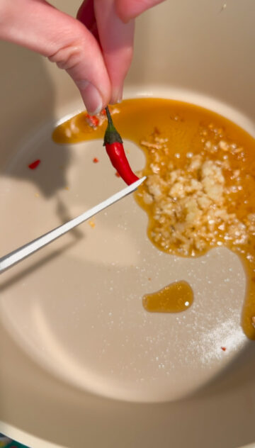 Thai chili peppers being cut and dropped into the beige-colored pot with pressed garlic and sesame oil.