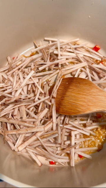 Julienned burdock roots and discs of Thai chili pepper being stir-fried in a beige-color pot.