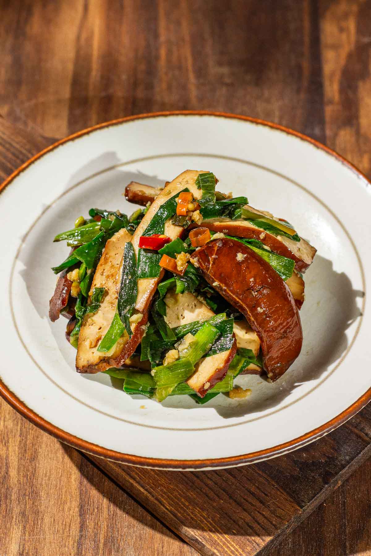 A plate of Chinese Garlic Chives Bean Curd Stir Fry, with bits of chopped red chili peppers and minced garlic on top, served in a white dish with brown rim on a wooden table.