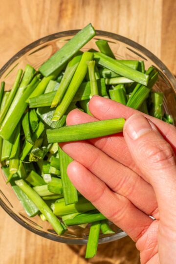 A hand holding a piece of chopped Chinese garlic chives, hovering over a bowl filled with similar pieces on a wooden surface.