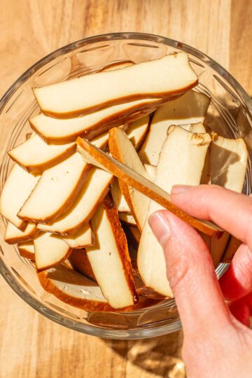 A hand holding a piece of bean curd slice, hovering over a bowl filled with similar slices on a wooden surface.