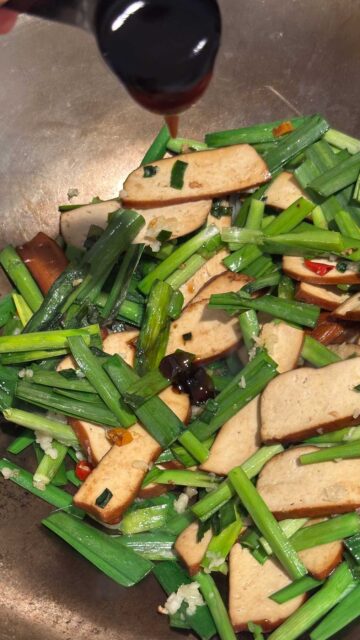 A spoonful of vegetarian oyster sauce being poured into a wok with bean curd slices and chopped Chinese garlic.