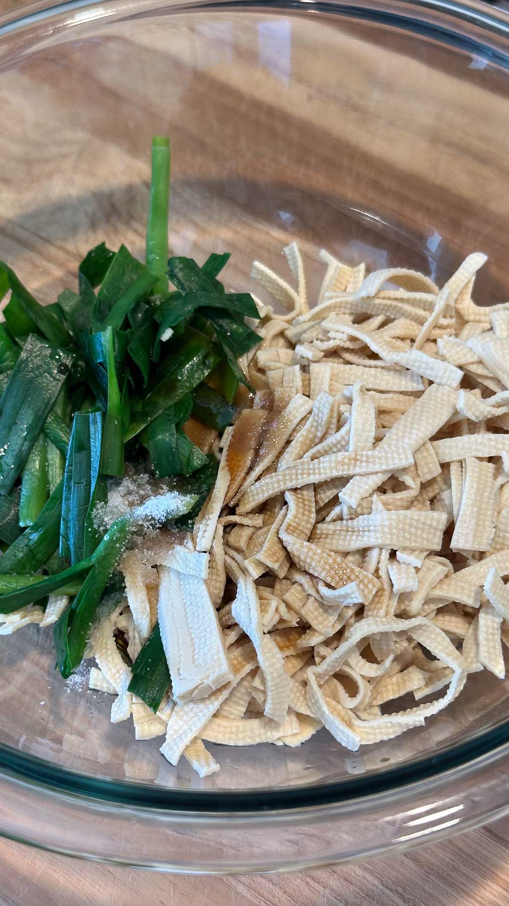 Blanched garlic chives and dried tofu noodles with seasonings in a clear glass bowl on a wooden surface.