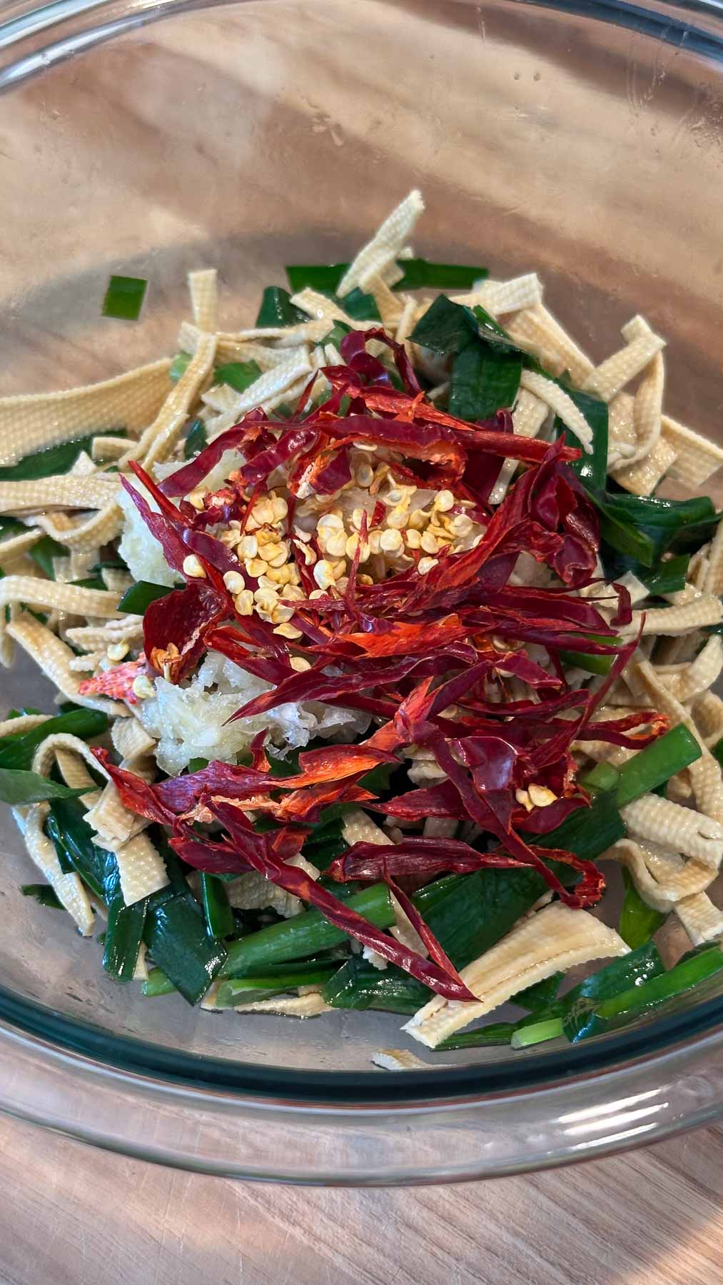 Minced garlic and dried chili pepper slices on top of blanched garlic chives and dried tofu noodles in a glass bowl on a wooden surface.