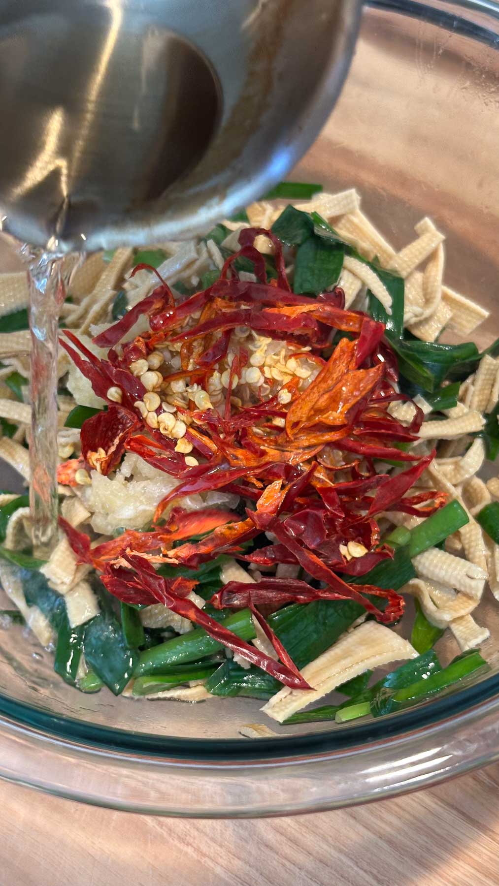 Hot oil being poured over dried chili pepper slices and garlic, on top of blanched garlic chives and dried tofu noodles in a glass bowl on a wooden surface.