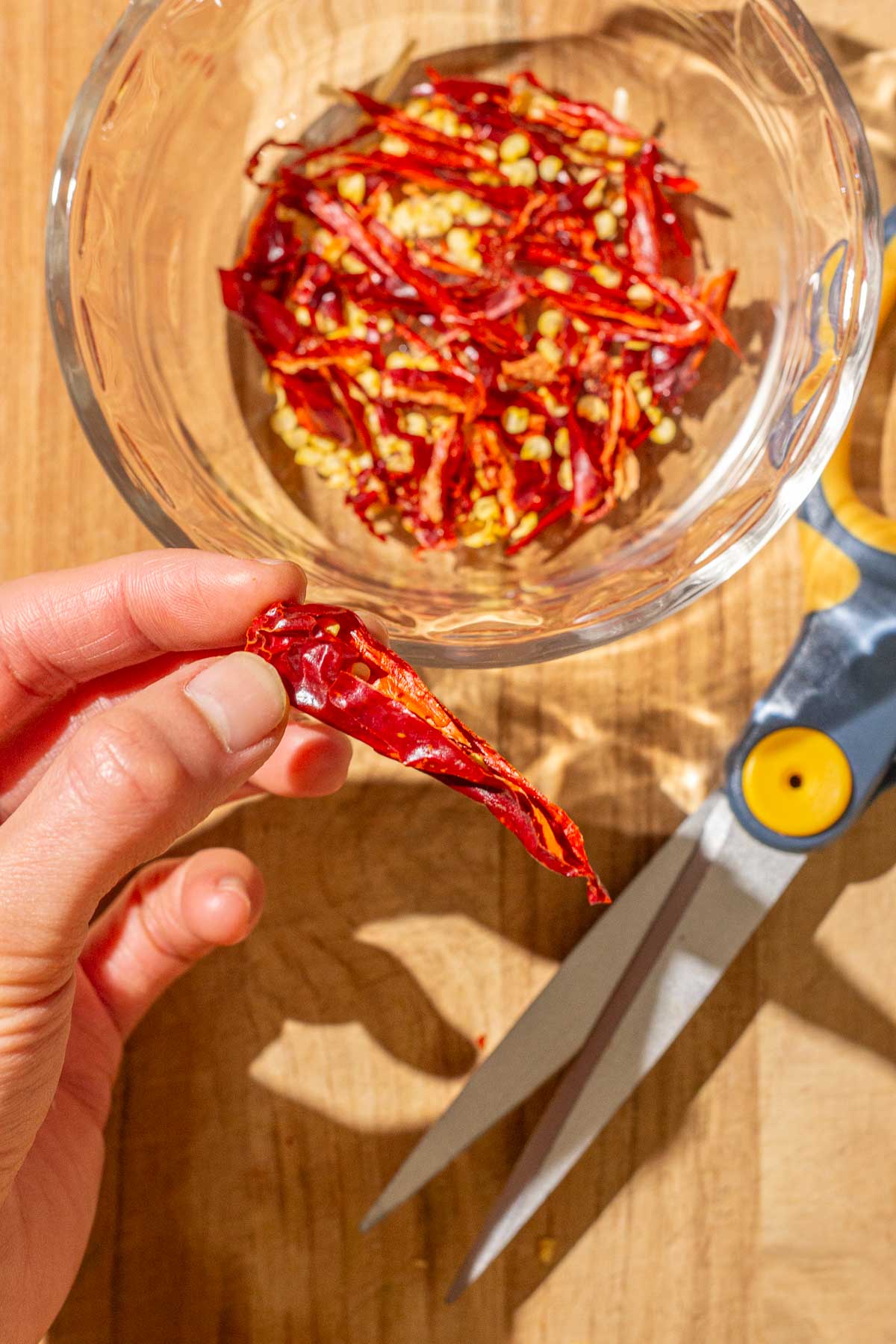 A hand holding a partially cut dried red chili pepper, above a bowl of chopped red chili peppers and a pair of scissors on a wooden surface.