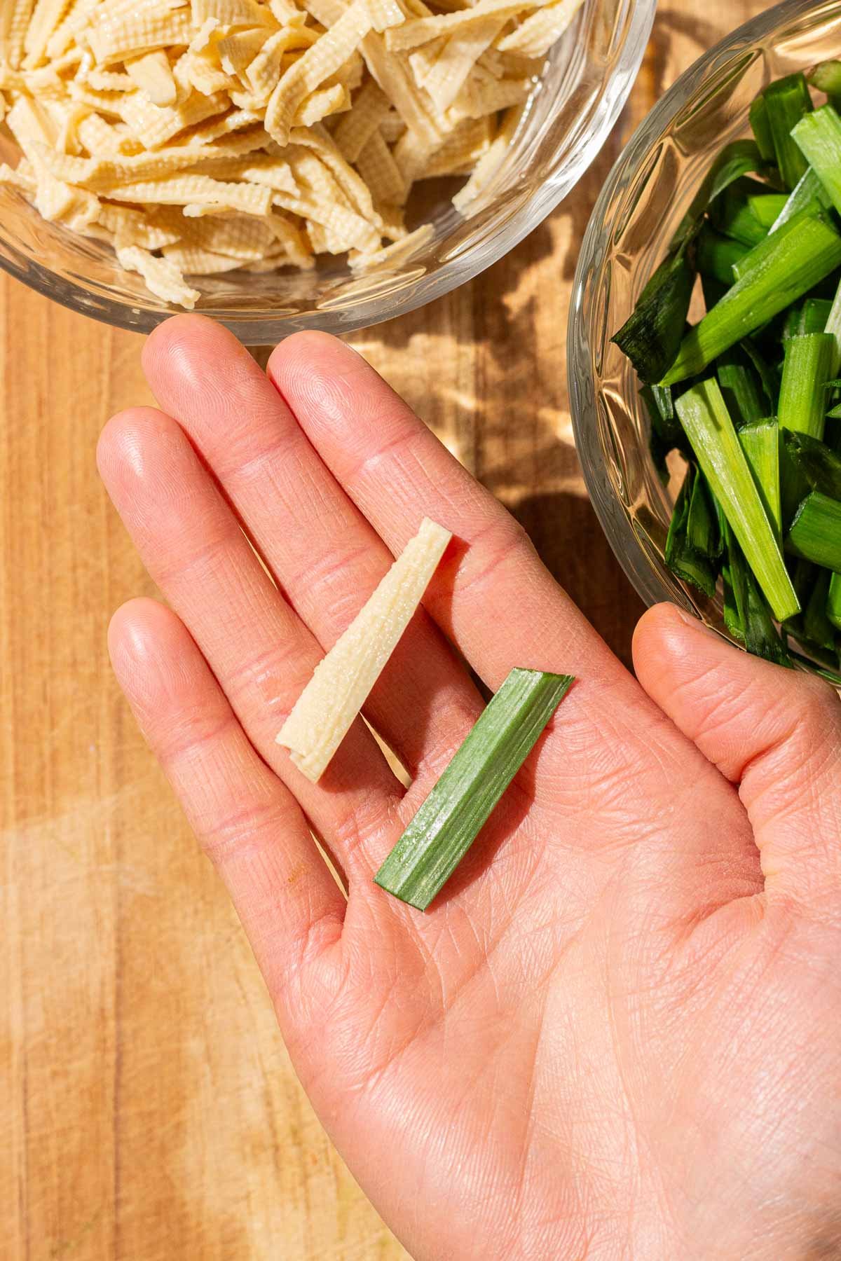 A hand holds one strip of dried tofu noodles and one strip of green garlic chive above bowls filled with ingredients for chives with dry tofu noodles.