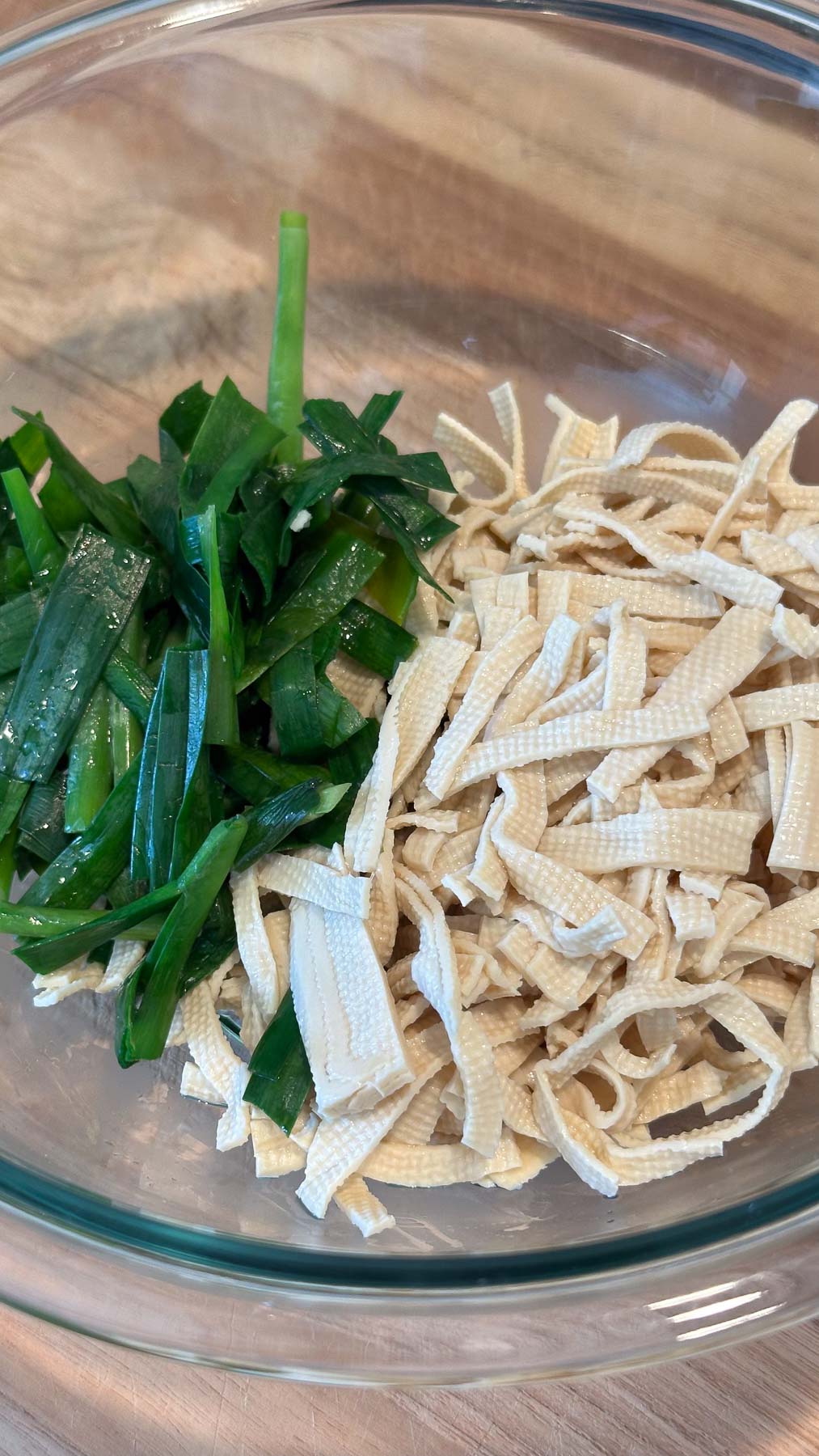 Blanched garlic chives and dried tofu noodles in a clear glass bowl on a wooden surface.
