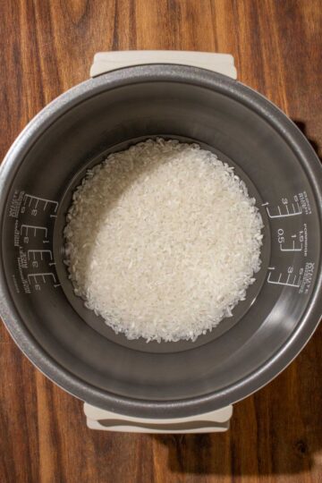 Uncooked white rice submerged in water in a rice cooker pot, set on a wooden surface.