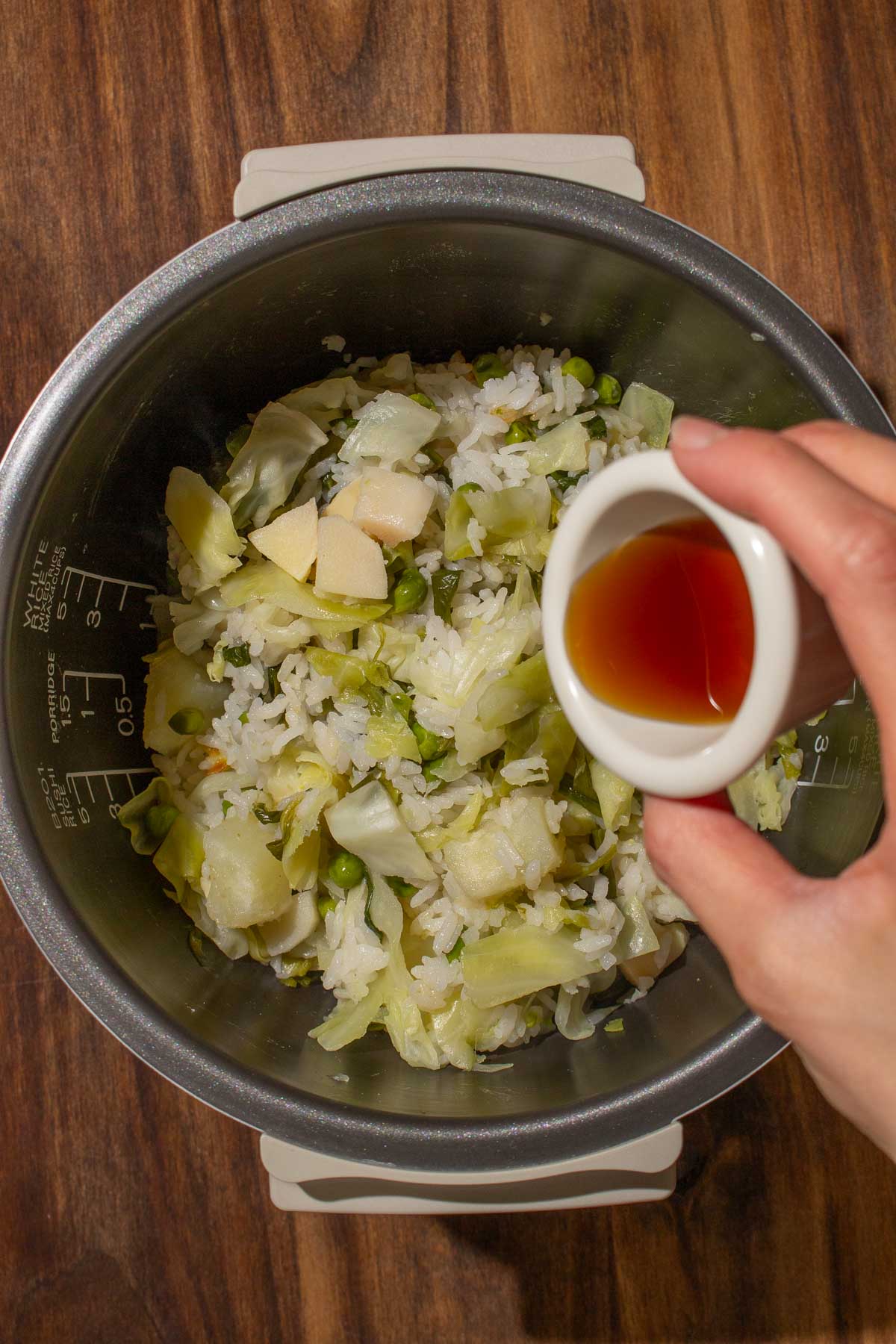 A hand pours sesame oil into a rice cooker pot with rice mixed with Chinese garlic chives, cabbage, peas, potatoes, and bamboo shoots.