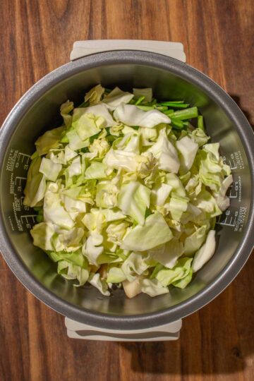 A bed of chopped and uncooked cabbage in a rice cooker pot on a wooden surface.