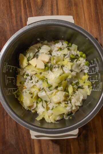 Rice mixed with Chinese garlic chives, cabbage, peas, potatoes, and bamboo shoots in a rice cooker pot on a wooden surface.