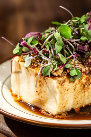 A block of silken tofu topped with a brown, savory sauce and microgreens, served on a white plate with a brown rim on a wooden table.