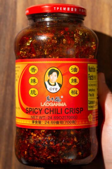 A hand holds a jar of Lao Gan Ma Spicy Chili Crisp, featuring a red label with Chinese text, a photo of the founder, partial nutrition facts, and visible chili oil and chili flakes inside.