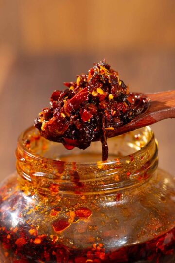 A close-up of a wooden spoon scooping chunky chili oil with red pepper flakes from a glass jar, with oil and spices visible on the jars rim.