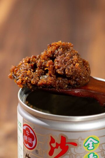 A close-up of a wooden spoon holding a chunky, brown paste over an open can of Bull Head Vegetarian Barbecue Sauce, with colorful Chinese characters on the label. The background is softly blurred.