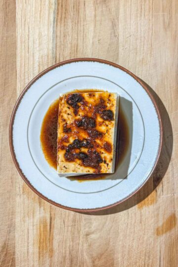 A block of silken tofu, topped with a dark, brown sauce, sits on a white plate on a wooden surface.