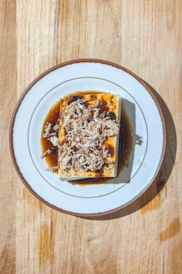 A block of silken tofu, topped with a dark, brown sauce and fried onions, sits on a white plate on a wooden surface.