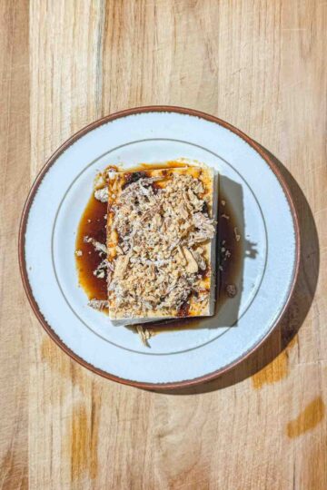 A block of silken tofu, topped with a dark, brown sauce, fried onions, and fried garlic, sits on a white plate on a wooden surface.