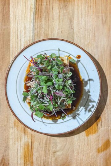 A block of silken tofu, topped with a dark, brown sauce, fried onions, fried garlic, and microgreens, sits on a white plate on a wooden surface.