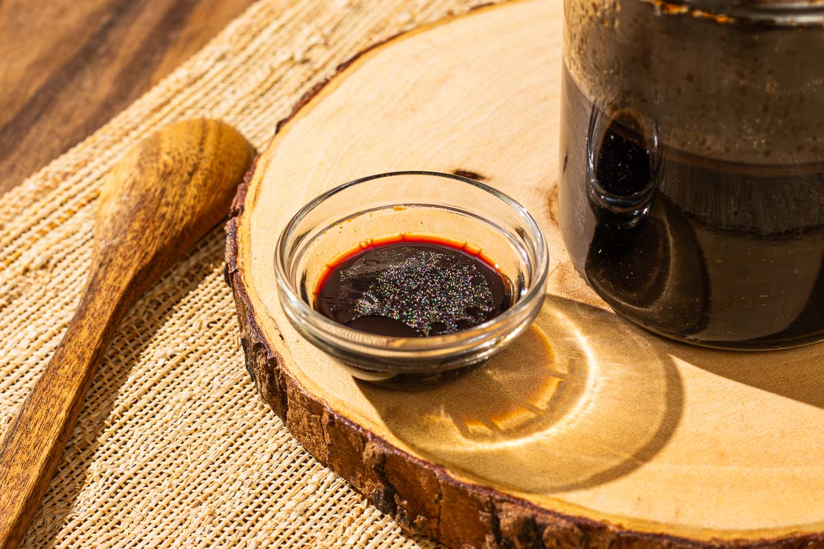 Small glass bowl of sweet soy sauce on a wooden tray, with a wooden spoon and jar nearby.