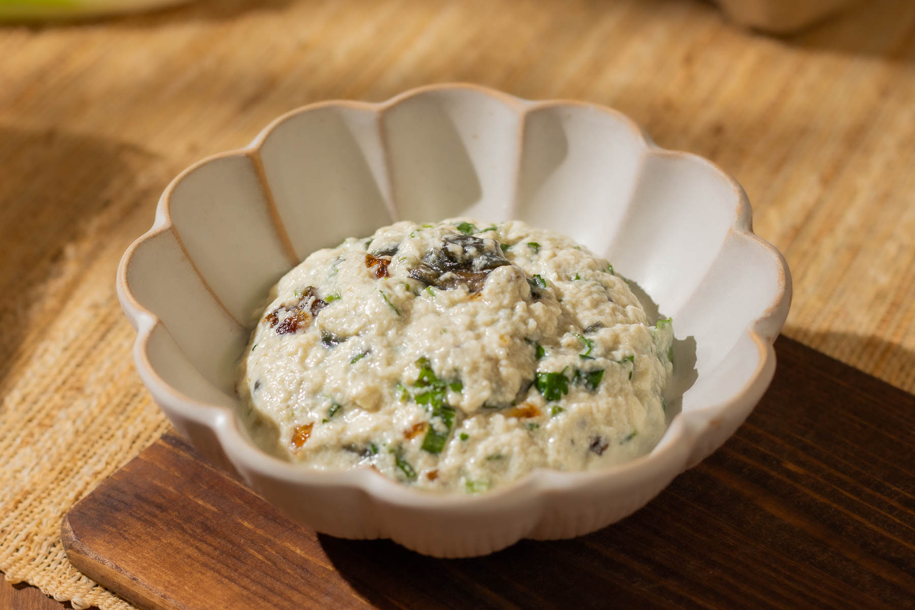A white scalloped bowl filled with mashed silken tofu, mixed with chopped century eggs and green onions, sits on a wooden board.