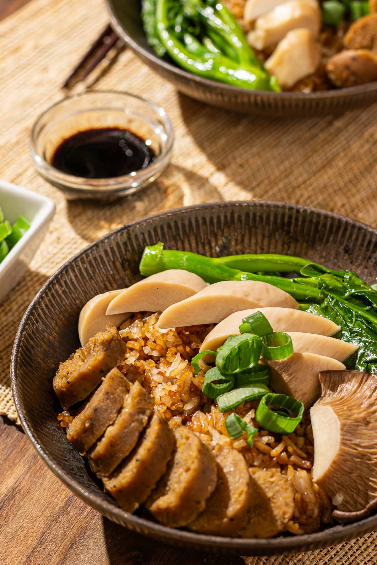 A bowl of rice cooker clay pot rice topped with sliced vegan sausage, mushrooms, gai lan, and green onions, with a small glass bowl of sweet soy sauce on the side.