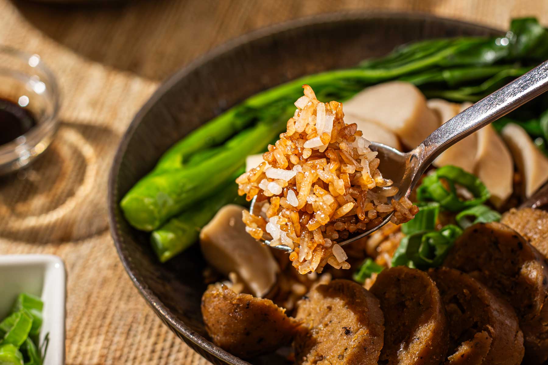 A spoonful of seasoned rice, above a bowl with vegetables and sliced vegan sausage.