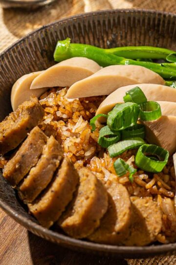 A bowl of rice cooker clay pot rice topped with sliced vegan sausage, mushrooms, gai lan, and green onions, with a small glass bowl of sweet soy sauce on the side.