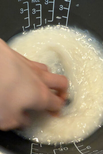 Hand stirring rice and water in a rice cooker pot, with measurement markings visible on the side.