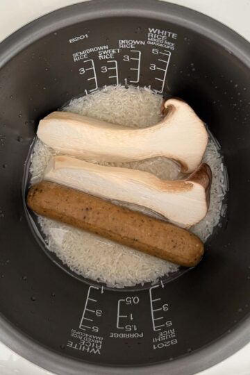 Uncooked white rice submerged in water in a rice cooker pot, with two king oyster mushroom halves and a sausage on top.