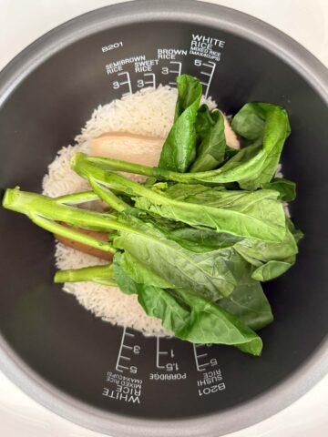 Uncooked gai lan on top of cooked white rice, king oyster mushrooms, and vegan sausage in a rice cooker pot with measurement markings.