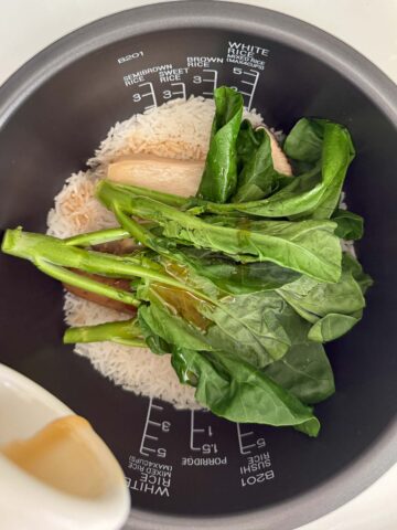 Uncooked gai lan on top of cooked white rice, king oyster mushrooms, and vegan sausage in a rice cooker pot, with sesame oil being poured in.
