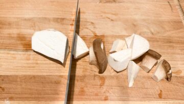 A knife cuts king oyster mushrooms into chunks on a wooden cutting board.