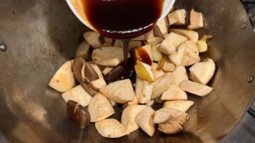 Chopped king oyster mushrooms cooking in a wok, with three cup sauce being poured over them from a bowl.