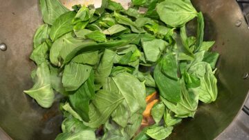 Fresh basil leaves cooking in a wok, with mushrooms partially visible underneath.